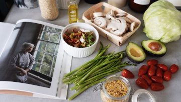A selection of fresh vegetables and a cookbook in a kitchen setting, promoting healthy cooking.